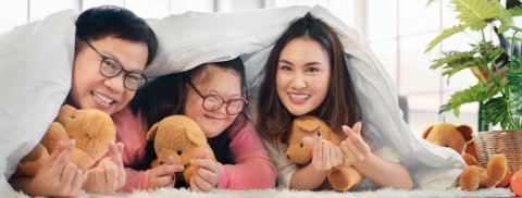 Asian parents smiling, play hiding under a blanket on the ground with their special needed daughter , all holding plushie bears.